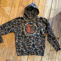 A camouflage hoodie with a brown and dark green camo pattern, featuring a central circular graphic with an orange boot and the text 'Everybody is cooler in cowboy boots, baby!. The hoodie is laid flat on a wooden plank floor.