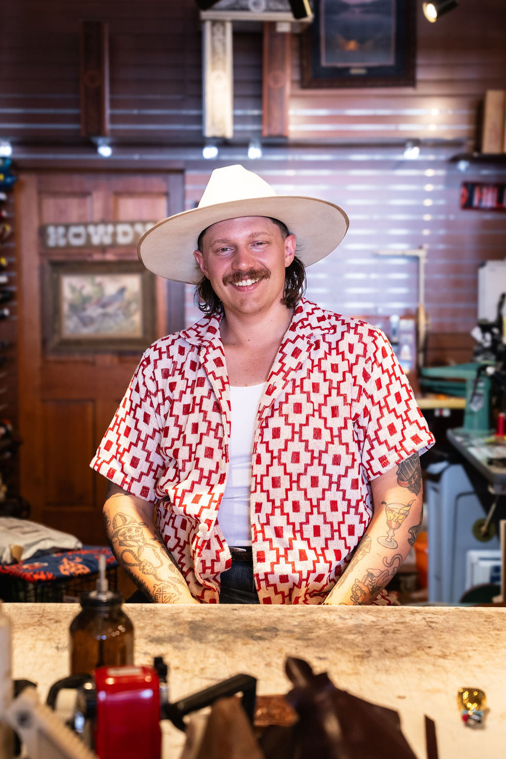 Jake the maker and owner wearing a white wide-brim hat, a red and white geometric patterned short-sleeve shirt, and a white undershirt stands behind a counter in his retail store