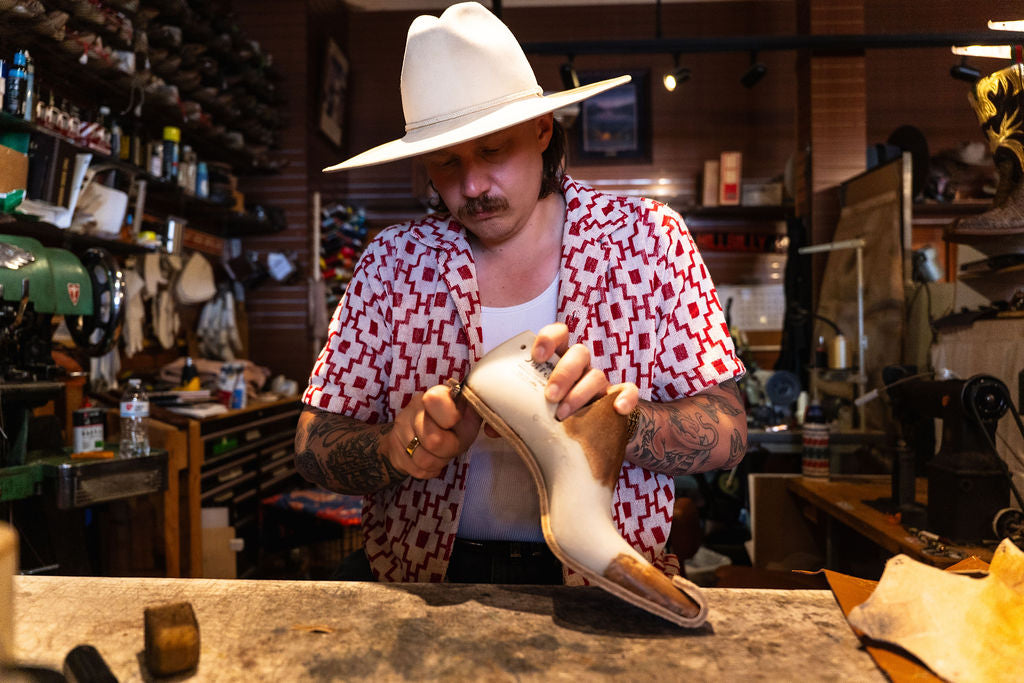 Jake in a white cowboy hat and a red-and-white patterned shirt working on a pair of boots in his workshop.