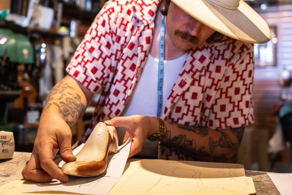 Jake the maker wearing a red and white patterned shirt and a wide-brimmed hat holds a beige leather shoe form on paper at a workbench in his workshop.