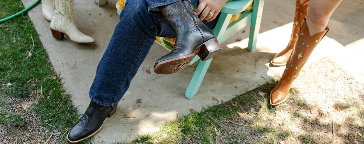 A person wearing blue jeans sits on a mint green chair outdoors, wearing black leather boots. Another person standing nearby wears tan western boots with white star-like cutouts.