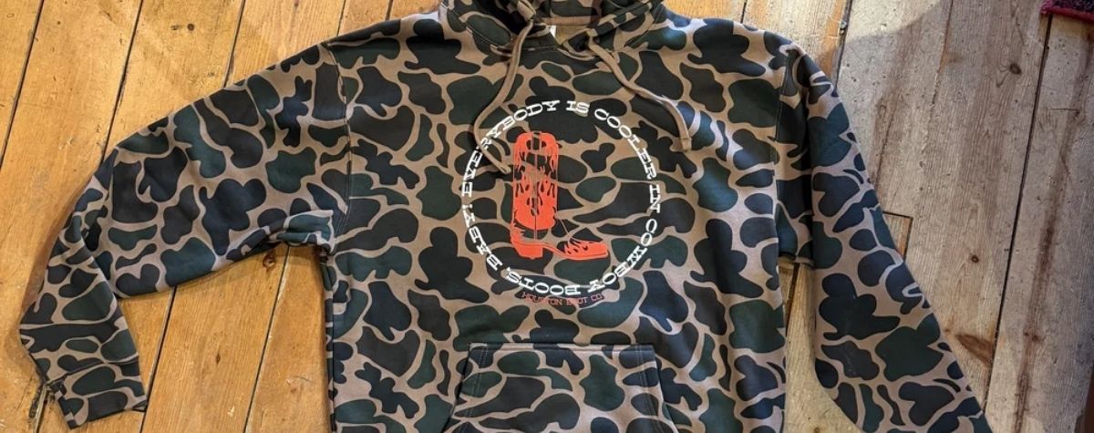A camouflage hoodie with a central circular graphic featuring a red boot and  text  laid flat on a wooden floor.